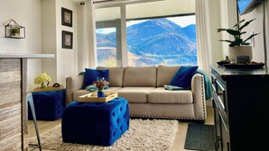 A living room with a sofa, ottoman, and a view of mountains through large windows. - 
Oasis @ Black Mountain - Only 25 mins to Big White (Kelowna)
