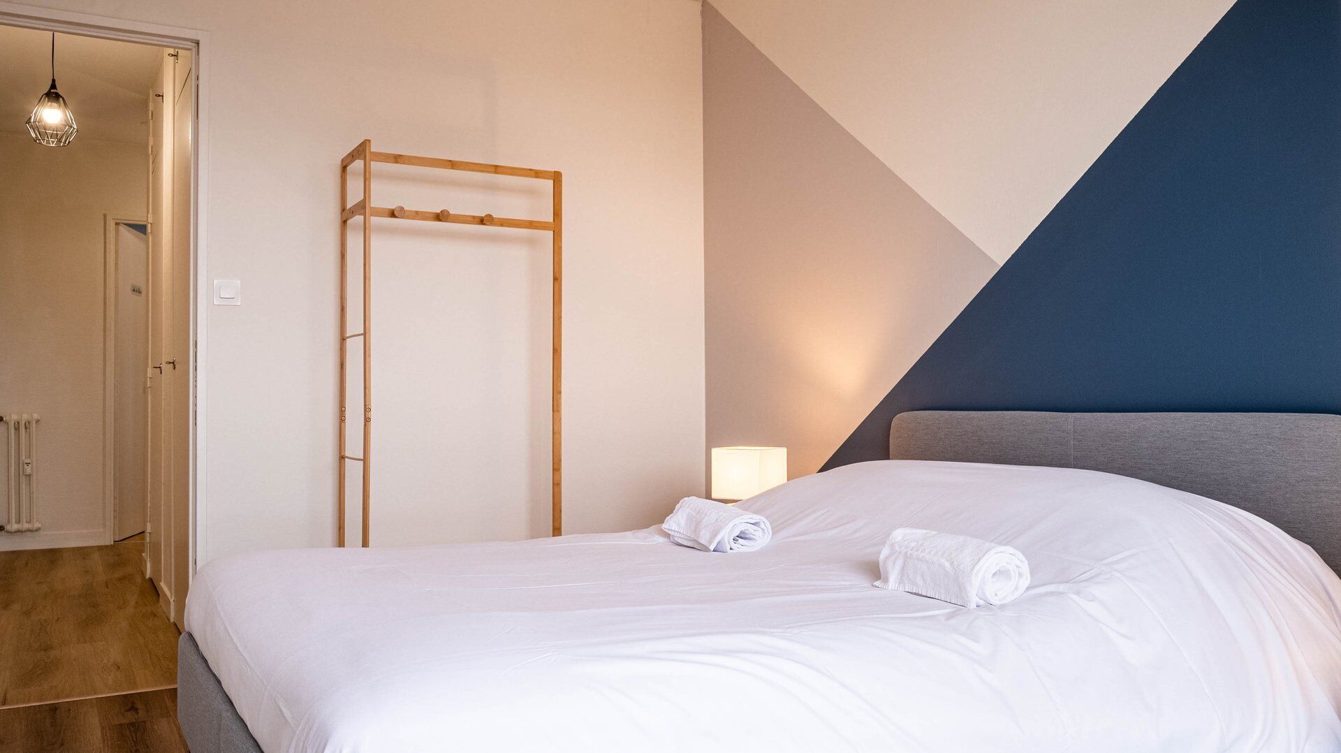 Ein ordentlich bezogenes Bett mit weißen Leinen, eine Holzleiter und eine Nachttischlampe in einem modernen Zimmer mit geometrischem Wanddekor.