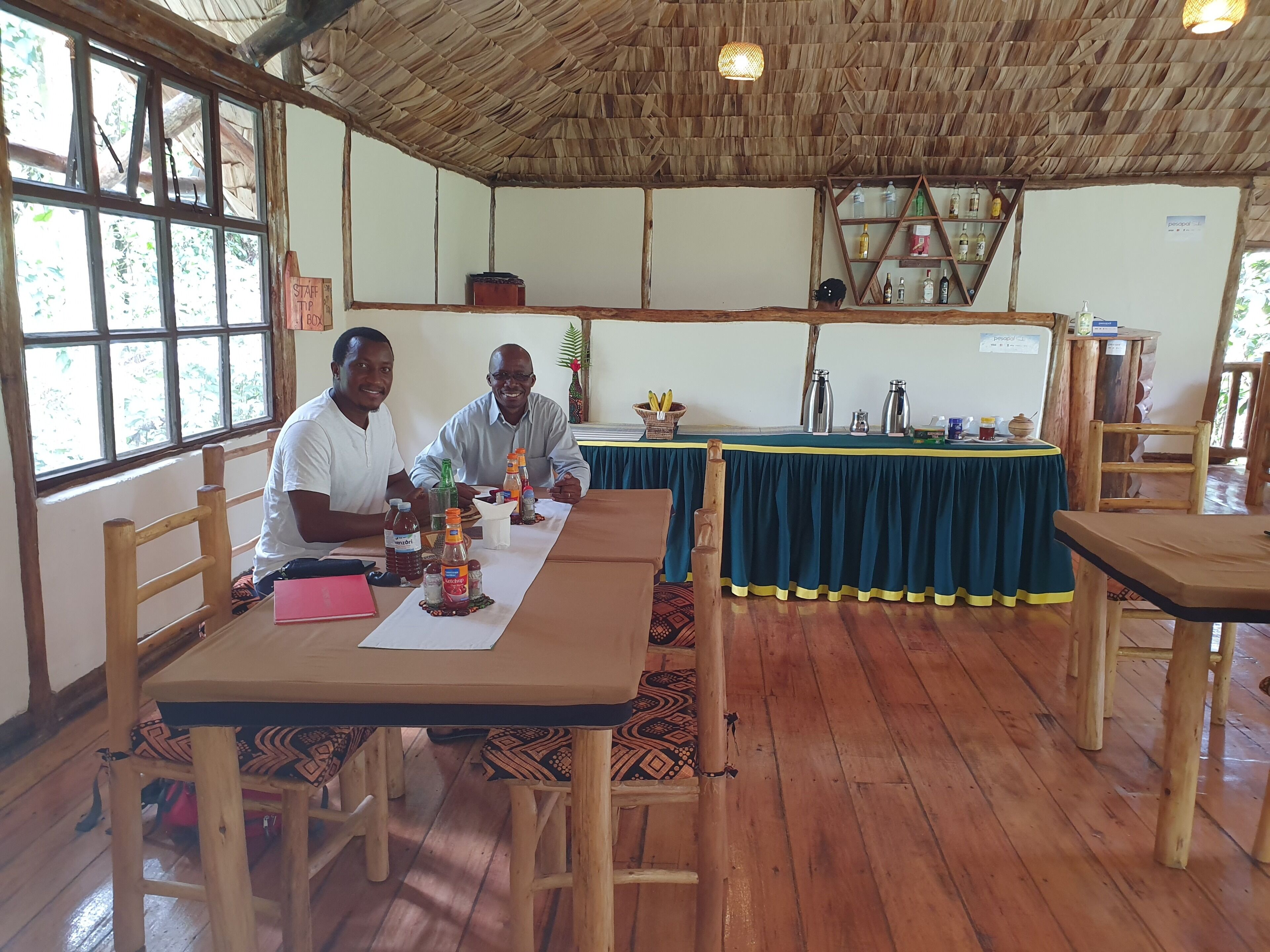 A rustic dining area with wooden tables and chairs, a thatched roof, and a view of greenery outside.