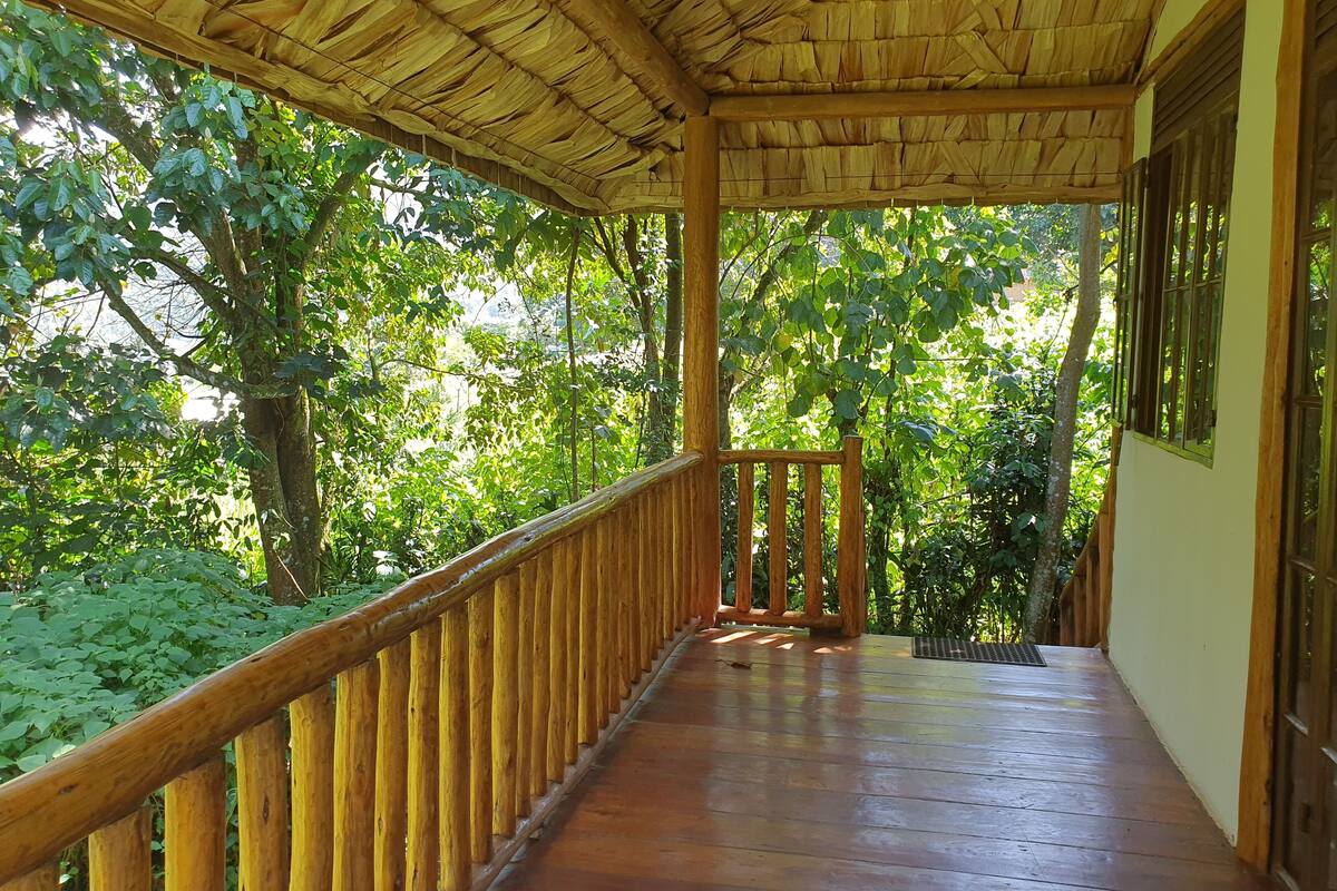A wooden balcony with a thatched roof, surrounded by lush greenery.
