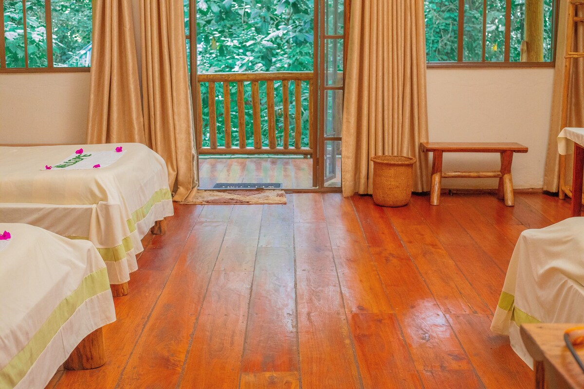 A room with wooden flooring, two beds with white linens, a wooden bench, and a view of greenery through a window.