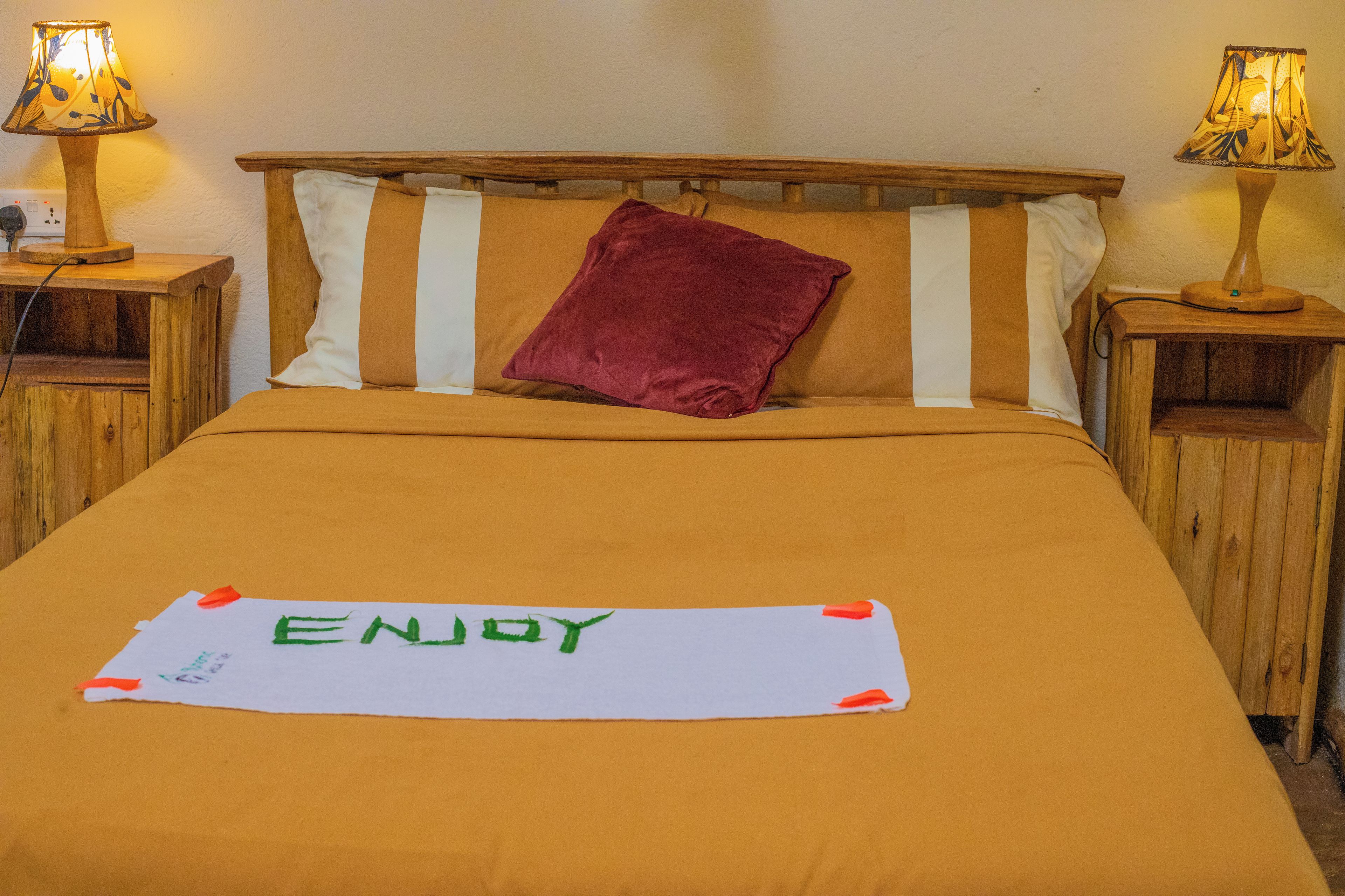 Ein Bett mit einer braunen Bettdecke, einem roten Kopfkissen und einem Schild mit der Aufschrift 'ENJOY'. Auf jeder Seite des Bettes befinden sich zwei Nachttische mit Lampen.