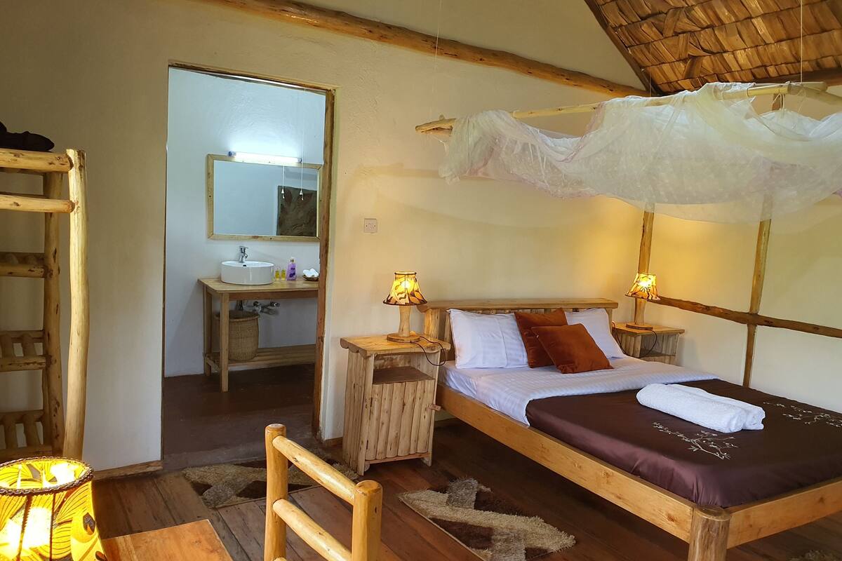 A wooden bed with a canopy, bedside lamps, a wooden chair, a small table, and a bathroom with a sink and mirror.