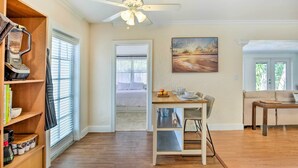A spacious living area with a wooden floor, a ceiling fan, a dining table with chairs, a sofa, and a bookshelf with books and a coffee maker.