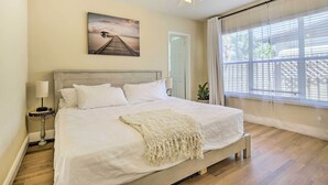 A bedroom with a large bed, a ceiling fan, a painting on the wall, and a window with blinds.