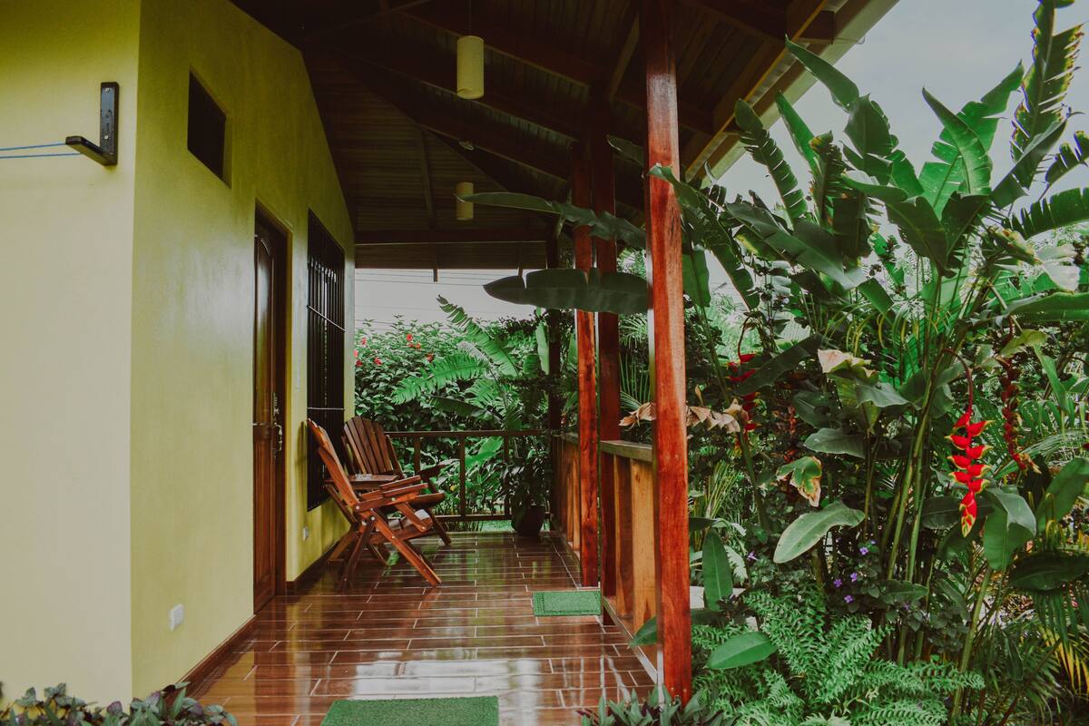 A covered porch with wooden chairs, a potted plant, and lush greenery.