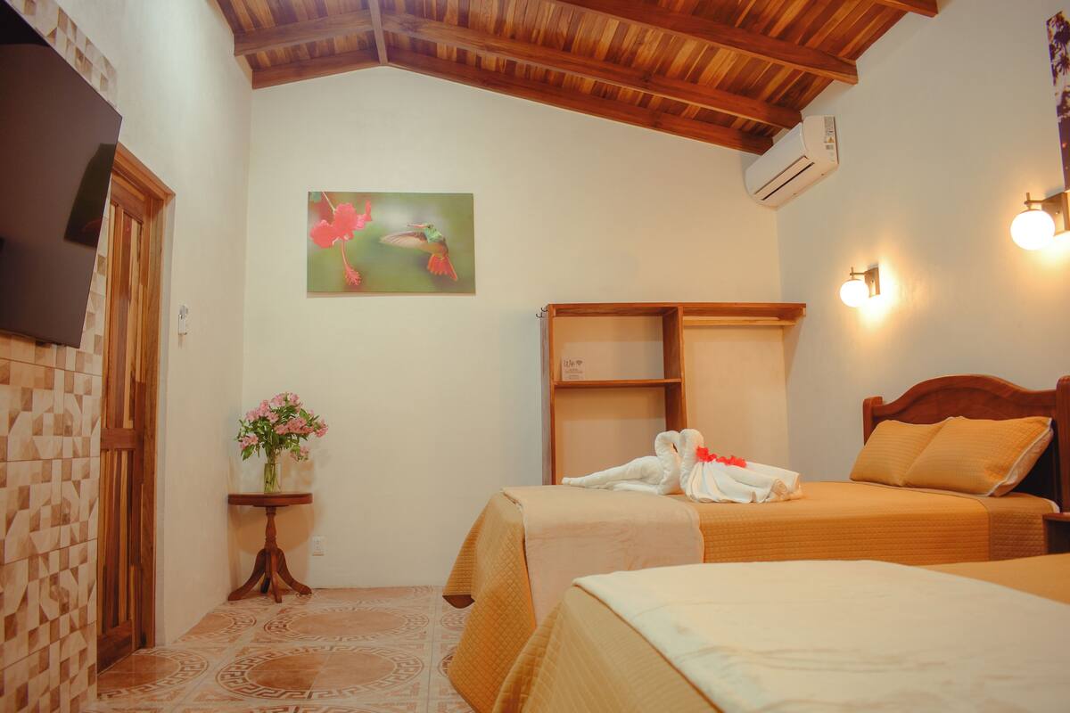 A hotel room with two beds, a wooden ceiling, a painting of birds, a small table with flowers, and a wall-mounted air conditioner.