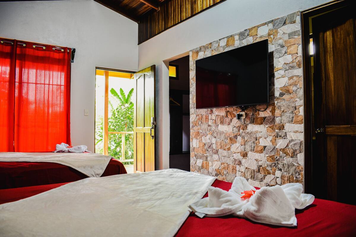 A hotel room with two beds, a stone wall, a TV, and a view of greenery outside.