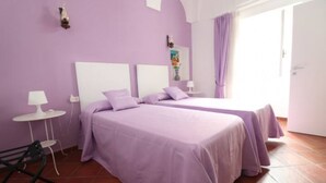 A room with two beds, each with a white headboard and purple bedding. There is a small round table with a lamp on one side of the room.