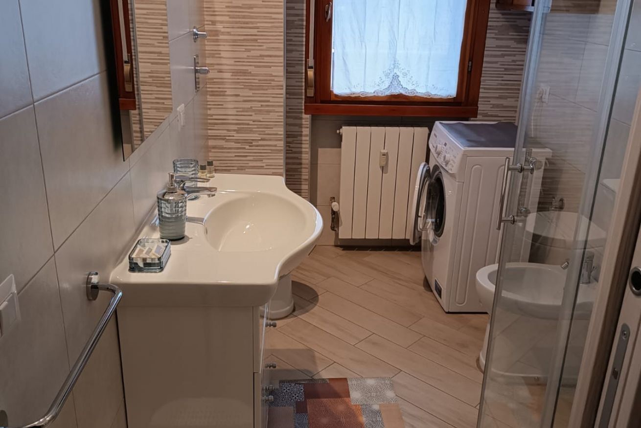 Een badkamer met wastafel, spiegel, raam, wasmachine en douchegedeelte.