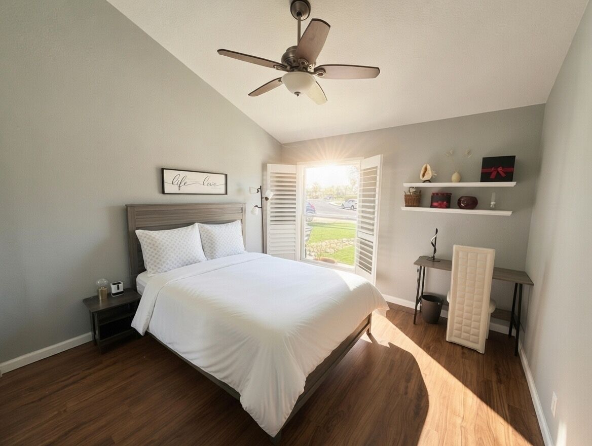 A bedroom with a bed, a ceiling fan, a nightstand, a window with shutters, and a shelf with decorative items.
