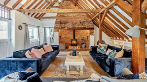 A living room with a brick fireplace, wooden beams, and two dark sofas. - Court Lodge is a substantial and picturesque twin roundel oast house set in pretty gardens within th (Heathfield)