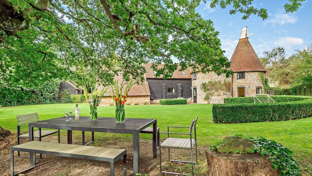 A rustic outdoor dining area with a table and chairs, surrounded by a well-maintained lawn and a traditional building with a conical roof. - Court Lodge is a substantial and picturesque twin roundel oast house set in pretty gardens within th (Heathfield)