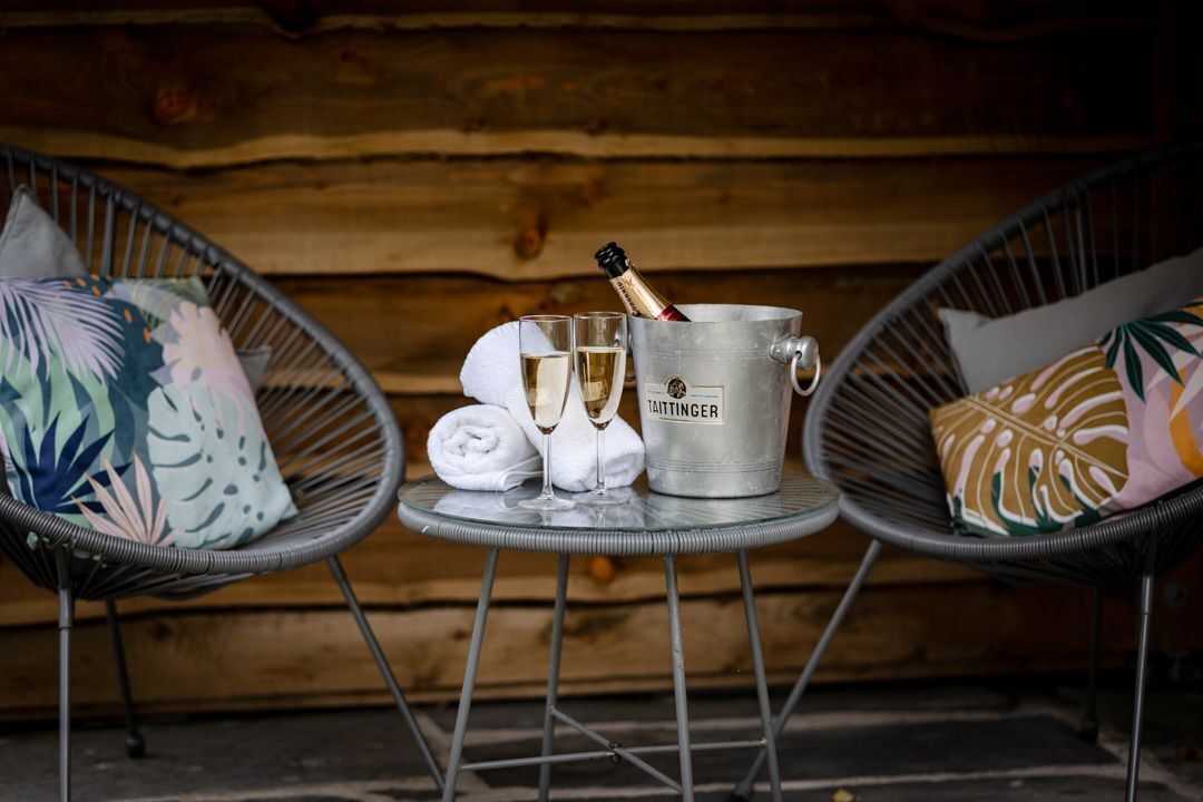 A cozy outdoor seating area with two wicker chairs, a metal table, a champagne bucket, a bottle of champagne, two glasses, and a towel.