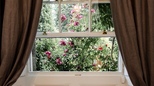 Un rincón acogedor junto a la ventana con cojines, una bandeja con tetera y un libro, con vistas a un jardín lleno de flores rosas.