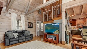 A spacious living room with a large flat-screen TV, a leather sofa, wooden floors, and a staircase leading to an upper level. - Enjoy cookouts on the deck and peaceful valley views! - Elemental Bliss (Jasper)
