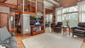 A spacious living room with a wooden staircase, a large flat-screen TV, leather sofas, and a dining area with natural light from large windows. - Enjoy cookouts on the deck and peaceful valley views! - Elemental Bliss (Jasper)