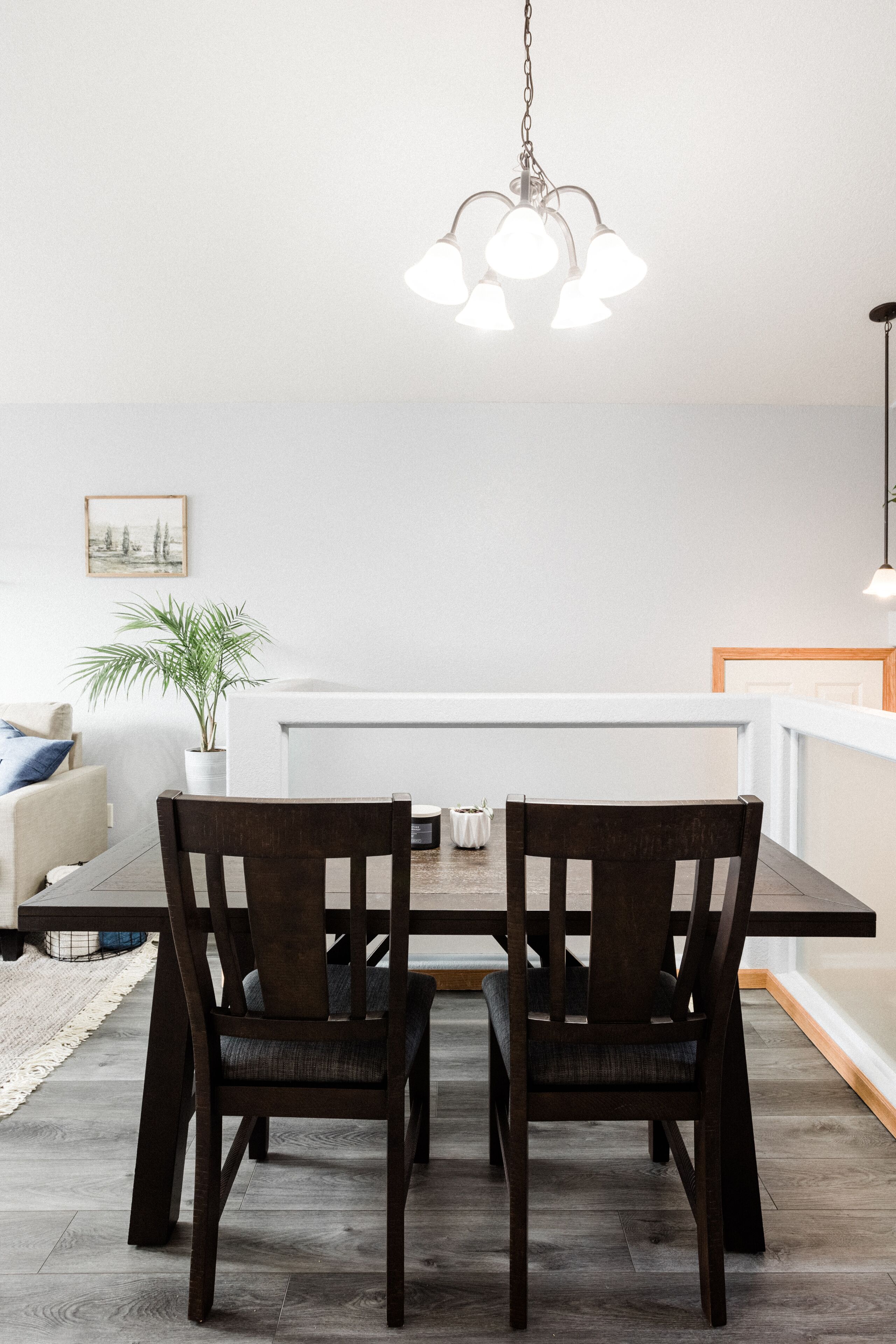 A dining area with a wooden table and chairs, a hanging light fixture, a potted plant, and a framed picture on the wall.