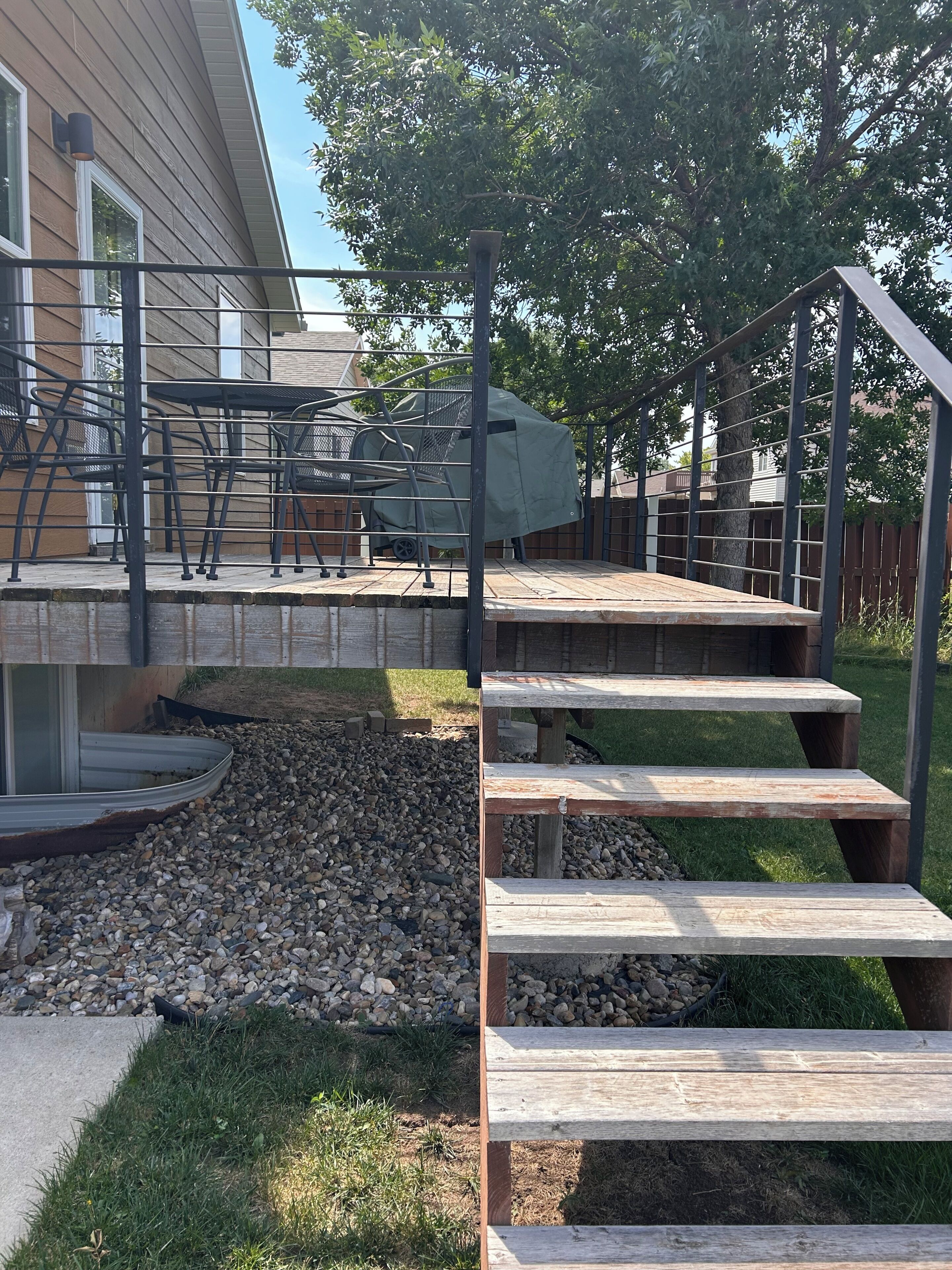 A wooden deck with outdoor furniture, a covered grill, and a staircase leading to a house.
