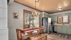 A rustic cabin interior with a wooden dining table, benches, a chandelier, and a kitchen with green cabinets and a refrigerator.
