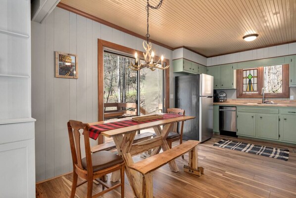 A rustic cabin interior with a wooden dining table, benches, a chandelier, and a kitchen with green cabinets and a refrigerator.