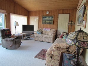 A cozy living room with wooden walls, a flat-screen TV, two sofas with floral upholstery, a lamp, and a painting on the wall. - Gorgeous and private hillside view overlooking Maple Bay (Duncan)