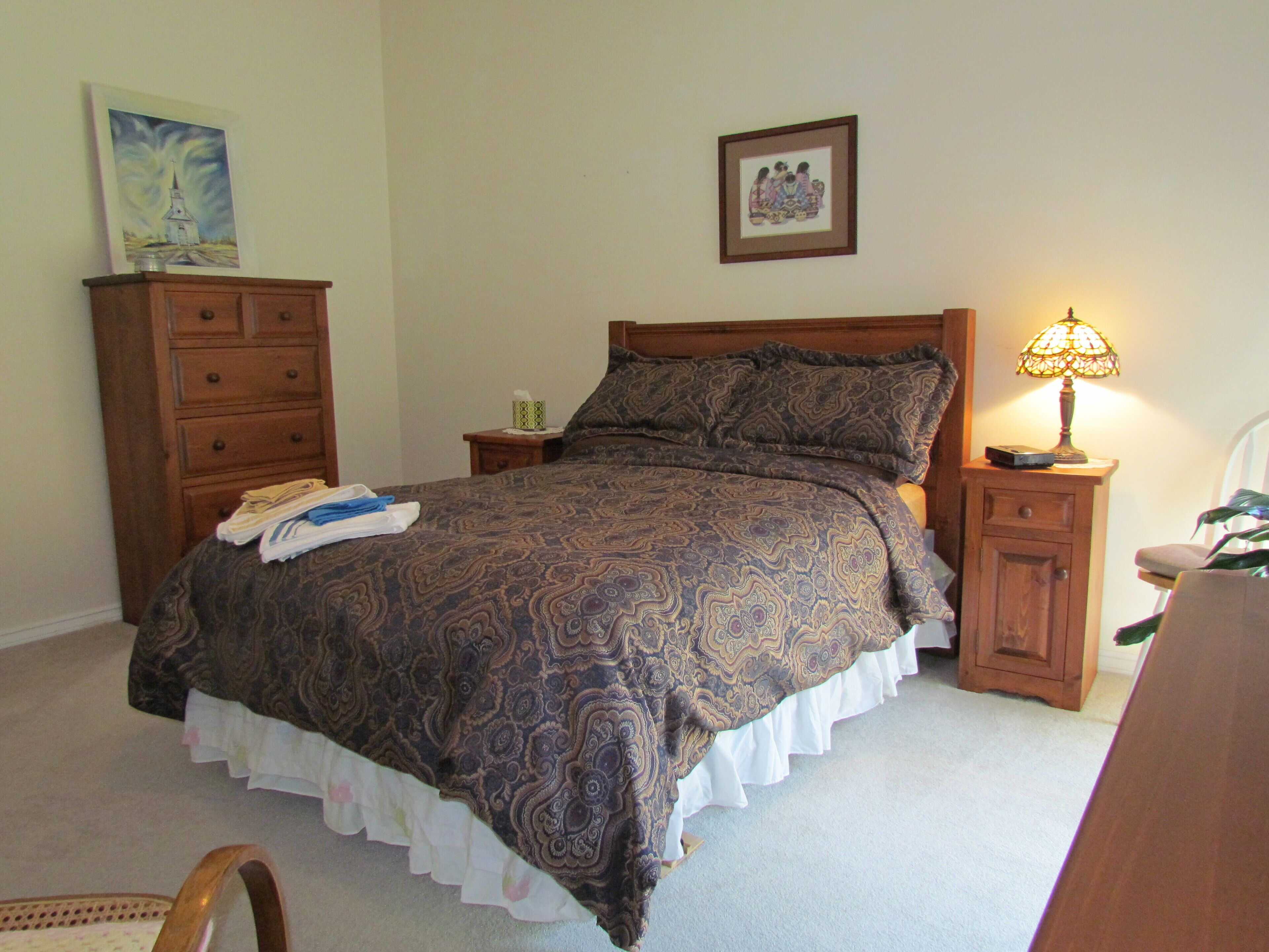 A bedroom with a bed, wooden nightstands, a lamp, a dresser, and two framed pictures on the wall.