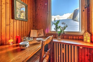 Interior de madera con vista a montañas nevadas, una mesa con una vela, una taza y un cuadro enmarcado.
