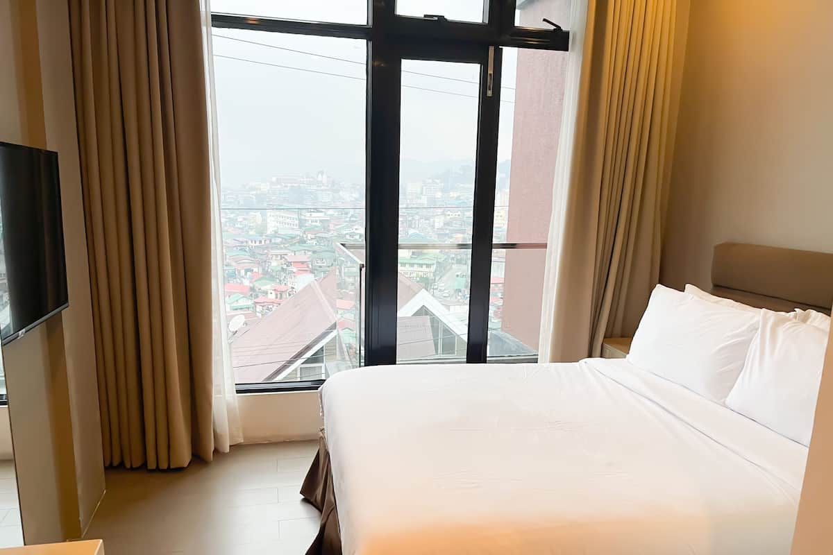 Habitación de hotel con una cama grande, un televisor y vistas a un paisaje urbano a través de un amplio ventanal.