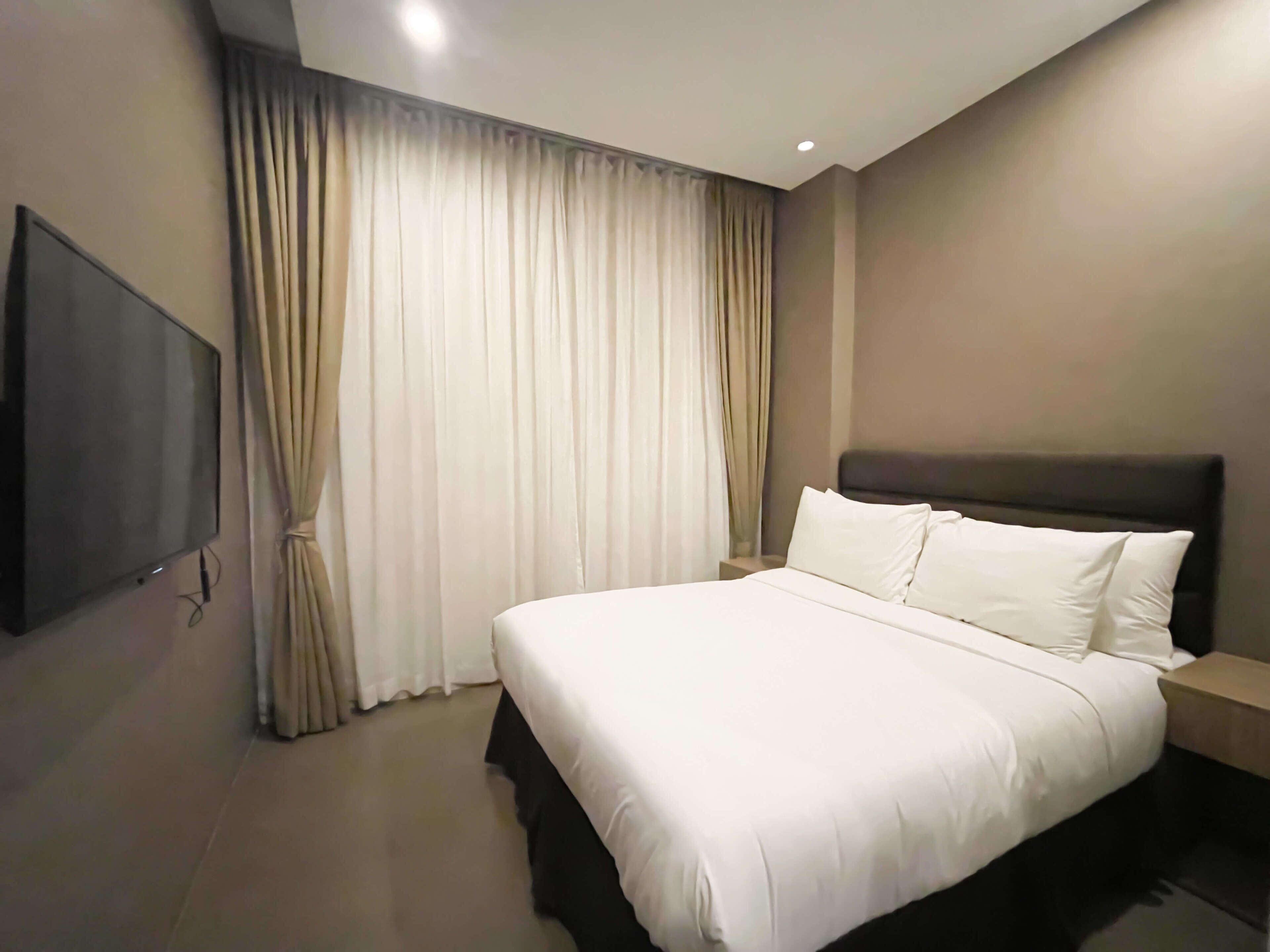A hotel room with a large bed, a flat-screen TV mounted on the wall, and a window with curtains.