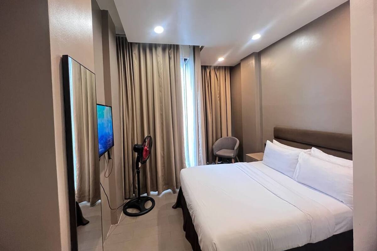 Una habitación de hotel con una cama grande, un televisor, un ventilador y una silla.