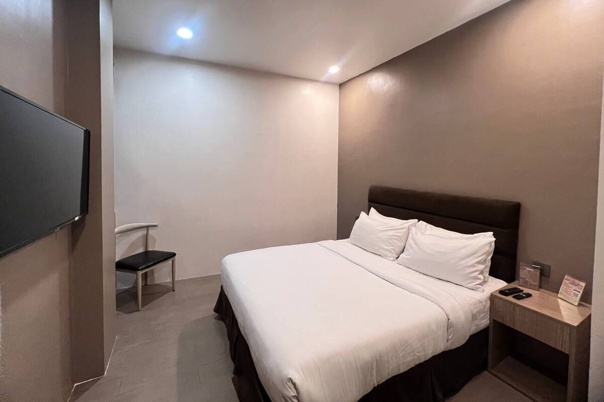 Una habitación de hotel moderna con una cama grande, un televisor de pantalla plana montado en la pared, una mesita de noche con lámpara y una silla.
