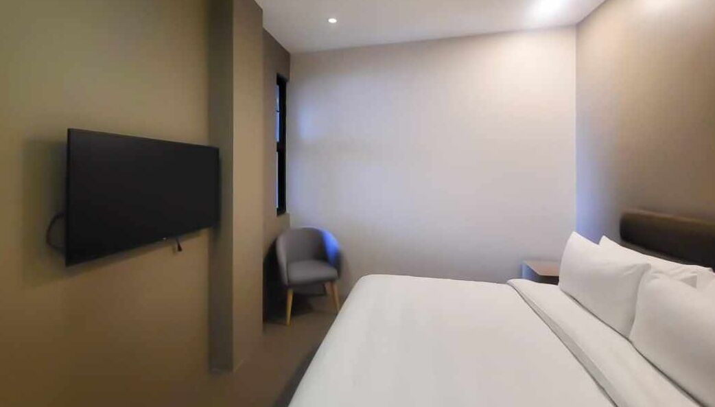 Une chambre d’hôtel avec un lit, une chaise, un téléviseur fixé au mur et une table de chevet.
