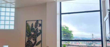 Una habitación amplia con un gran ventanal, un sofá rojo, un sofá verde, una mesita redonda y una lámpara en el techo.