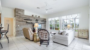 A spacious living room with a white sofa, a black chair, a coffee table, and a TV. There is a wooden accent wall with shelves and a ceiling fan. - Solar Heated Pool, Hot Tub, Kayaks, Canal Views - Villa Tranquil Haven- Roelens Vacations (Cape Coral)