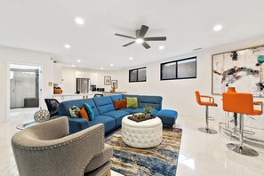 A modern living room with a blue sofa, a grey armchair, and a white ottoman. There is a dining area with orange chairs and a glass table. The room has a ceiling fan and recessed lighting.