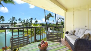 A balcony with a wicker sofa, a dining table with a fruit bowl, and a view of a lake with palm trees.