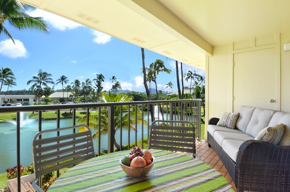 A balcony with a wicker sofa, a dining table with a fruit bowl, and a view of a lake with palm trees. - Kauai Beach Villas, Walk to beach, FREE PARKING (Lihue)