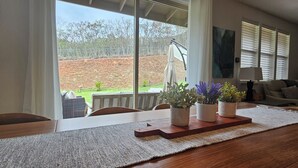 A dining area with a large window overlooking a fenced backyard. There is a wooden table with a placemat and two potted plants on it.