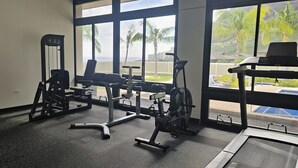 A gym with various exercise equipment, large windows with a view of palm trees and buildings.