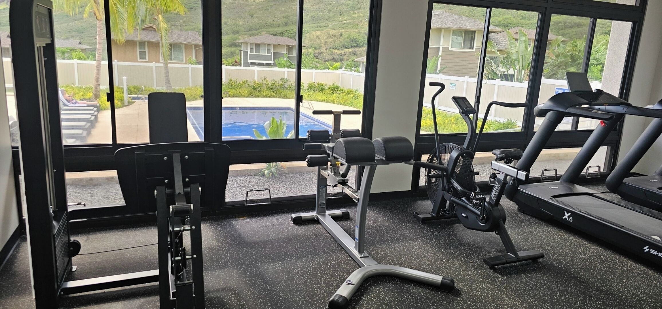 A gym with exercise equipment, a treadmill, and a view of a residential area through large windows.