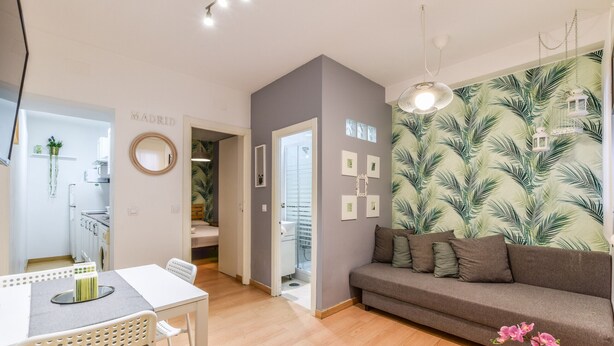 A living room with a grey sofa, a dining table with chairs, a kitchen area, and a bathroom visible through an open door. - apartamento en lavapies de alquiler temporal (Madrid)