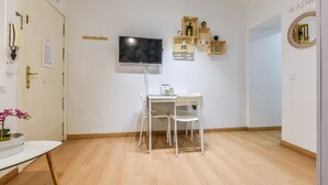 A room with a white desk, two white chairs, a flat-screen TV mounted on the wall, wooden shelves with decorative items, and a door with a nameplate. - apartamento en lavapies de alquiler temporal (Madrid)