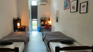 Una habitación estrecha con dos camas individuales, un escritorio con silla y una mesita con lámpara.