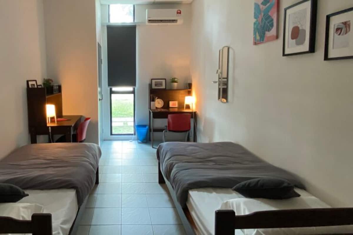 Quarto pequeno com duas camas de solteiro, uma escrivaninha com cadeira e uma mesinha de cabeceira com abajur.