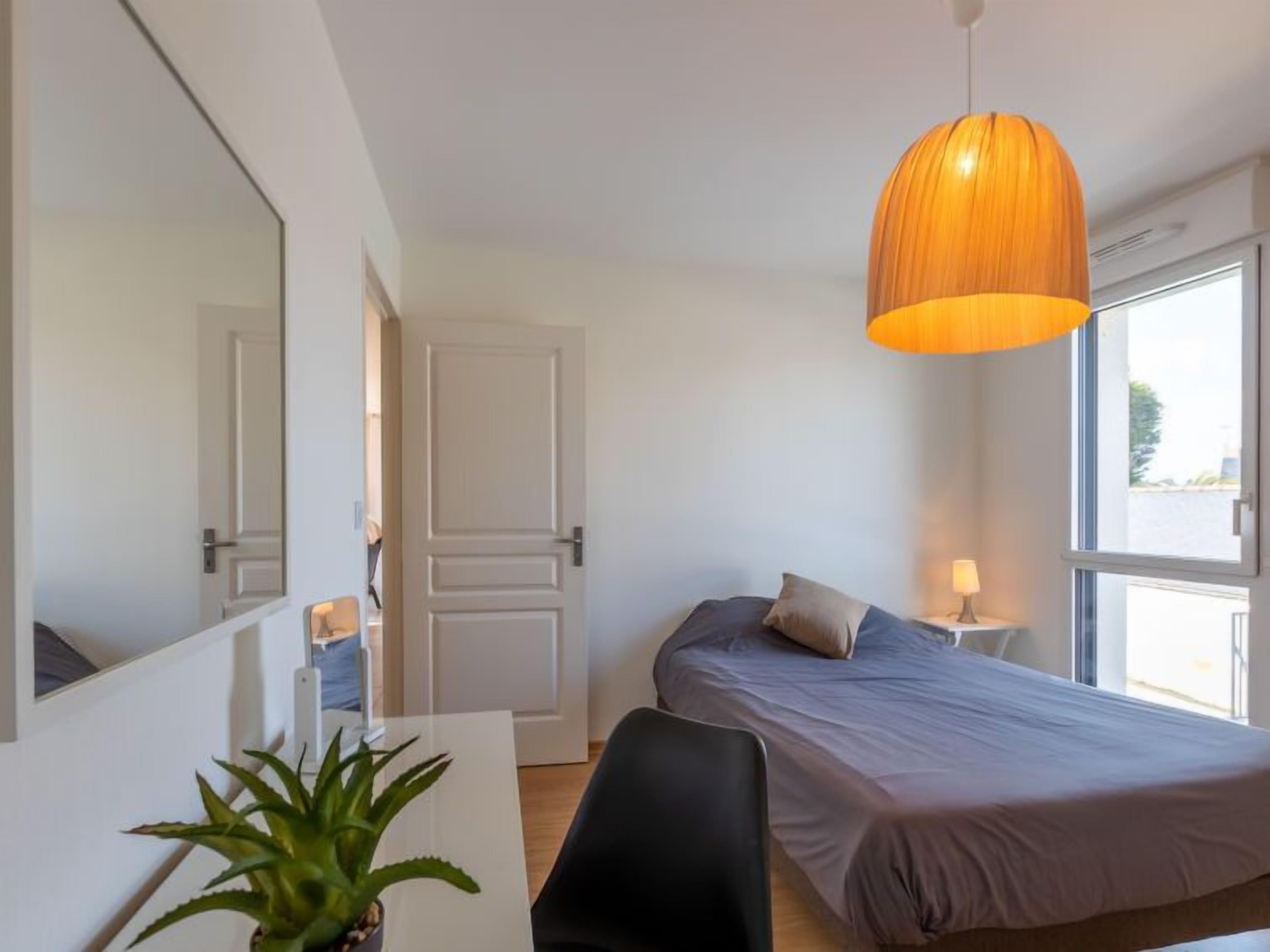 A bedroom with a bed, a desk, a chair, a potted plant, and a hanging light fixture.