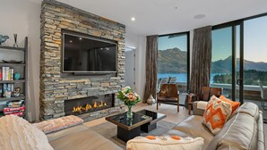 A modern living room with a stone fireplace, a flat-screen TV, comfortable seating, and a view of mountains through large windows. - Chic Central Penthouse & Spa by Staysouth (Queenstown)