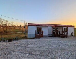 A small, single-story building with a red roof and white siding, featuring a porch with wooden railings and a gravel driveway. - Cowboy Cottage (Florence)