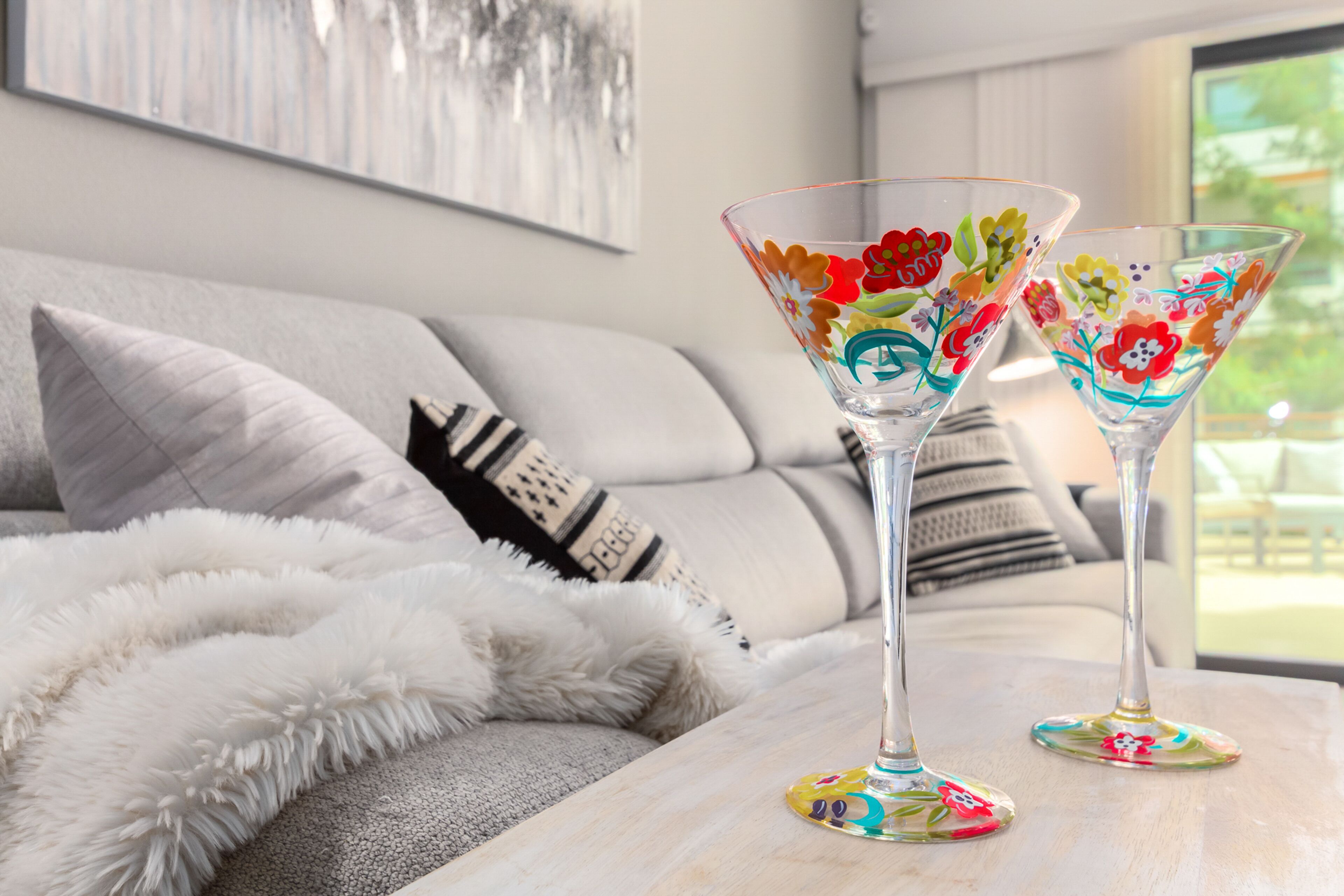 Two clear glass martini glasses with floral designs on a light-colored table in a modern living room with a sofa and a white furry throw blanket.