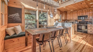 A wooden cabin interior with a dining area, kitchen, and a large window overlooking greenery. - Tranquility Ranch - Hot Tub | Pool Table | Fire Pit in Private Yard (Mineral Bluff)